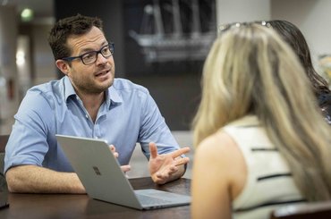 A man speaks to two women in front of a laptop
