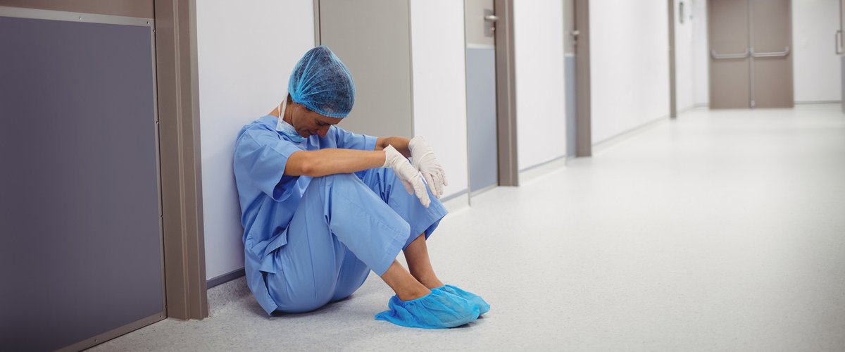 A tired surgeon sits on the floor of a hospital corridor with their head hanging down.