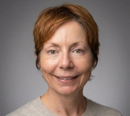 Head and shoulders portrait of a person with short auburn hair wearing a light-coloured jumper, photographed against a plain grey background.