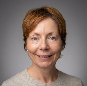 Head and shoulders portrait of a person with short auburn hair wearing a light-coloured jumper, photographed against a plain grey background.