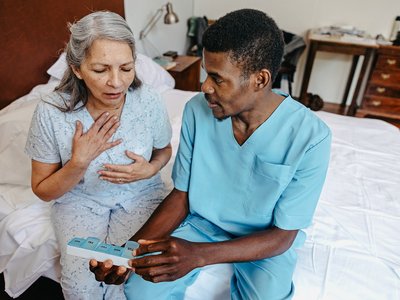 A person in bed sits beside a healthcare worker holding a weekly medication organiser. The person places a hand on their chest while the healthcare worker appears to discuss their medicines in a care home setting.