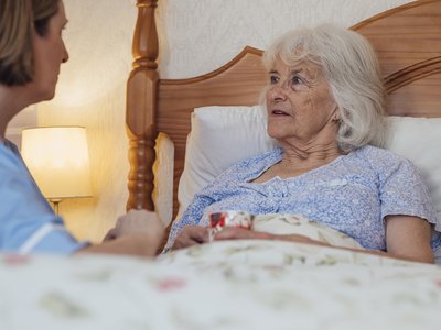 Older person in bed at home holding a mug and speaking with a carer seated beside the bed, with a bedside lamp and wooden headboard in the background.