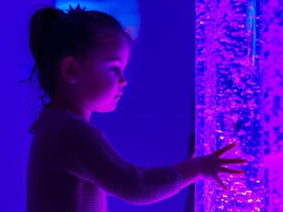 Child reaches towards a glowing bubble tube in a sensory room, illuminated by pink and purple lights. The tube is filled with rising bubbles and glowing beads, casting coloured light across the scene.
