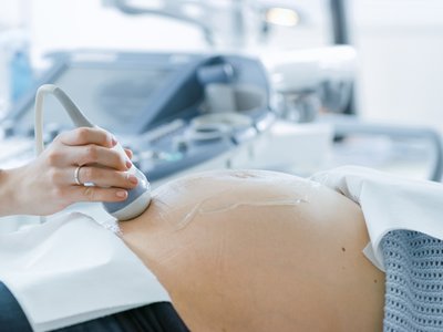 Close-up of an ultrasound scan being performed on a pregnant person's abdomen in a clinical setting, with the ultrasound probe held against the skin and medical equipment visible in the background.