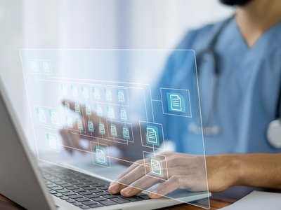 A doctor wearing blue scrubs uses a laptop. A graphic of computer folders to represent electronic patient record systems is superimposed on the photo.