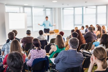 A mixed group of adults sit in rows in a bright classroom, listening to an educator deliver a course.