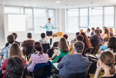 A mixed group of adults sit in rows in a bright classroom, listening to an educator deliver a course.