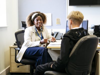 A healthcare professional sits in a consulting room speaking with a young person seated opposite. A desk, computer screens and office equipment are visible in the background.