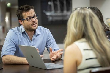 A man speaks to two women in front of a laptop