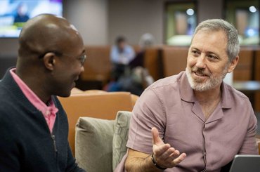 Two colleagues sit together in a lounge-style workspace, engaged in discussion. One person gestures while speaking as they sit beside a laptop, with seating areas and other people visible in the background.