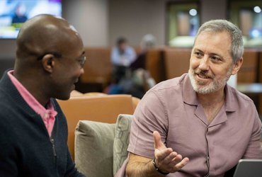 Two colleagues sit together in a lounge-style workspace, engaged in discussion. One person gestures while speaking as they sit beside a laptop, with seating areas and other people visible in the background.