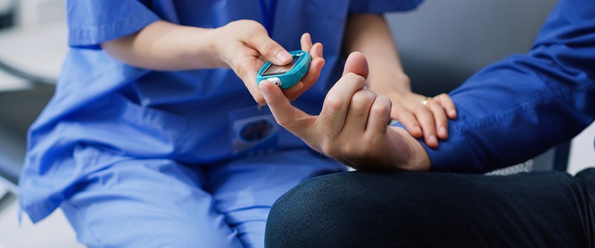 A healthcare assistant wearing blue scrubs checks the blood sugar level of a male patient.