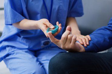 A healthcare assistant wearing blue scrubs checks the blood sugar level of a male patient.