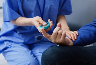 A healthcare assistant wearing blue scrubs checks the blood sugar level of a male patient.