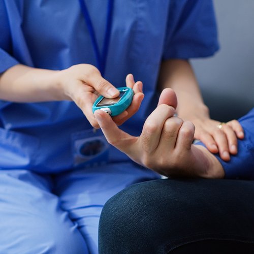 A healthcare assistant wearing blue scrubs checks the blood sugar level of a male patient.