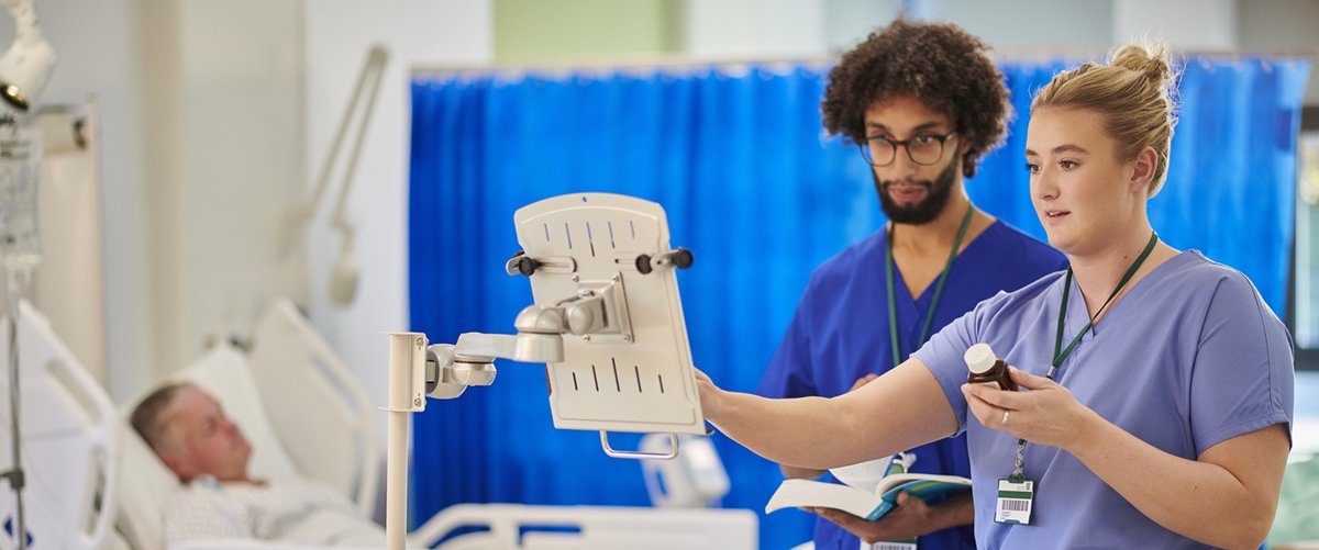 Healthcare staff prepare and check medicines on a hospital ward using a drugs trolley, following a digital written record beside a patient’s bed.