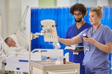Healthcare staff prepare and check medicines on a hospital ward using a drugs trolley, following a digital written record beside a patient’s bed.