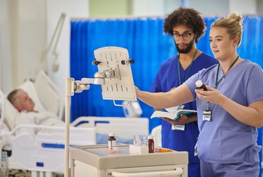 Healthcare staff prepare and check medicines on a hospital ward using a drugs trolley, following a digital written record beside a patient’s bed.