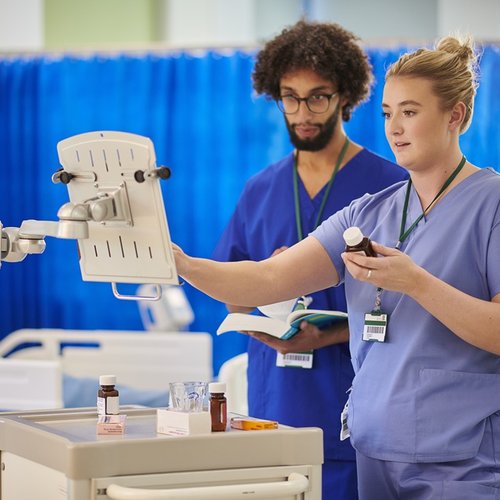 Healthcare staff prepare and check medicines on a hospital ward using a drugs trolley, following a digital written record beside a patient’s bed.