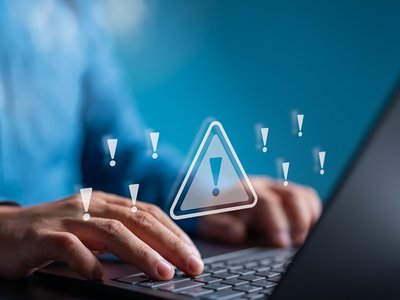 Hands type on a laptop keyboard. Nine exclamation marks are superimposed over the photo, including one in a warning triangle, to signify an issue with the computer system.