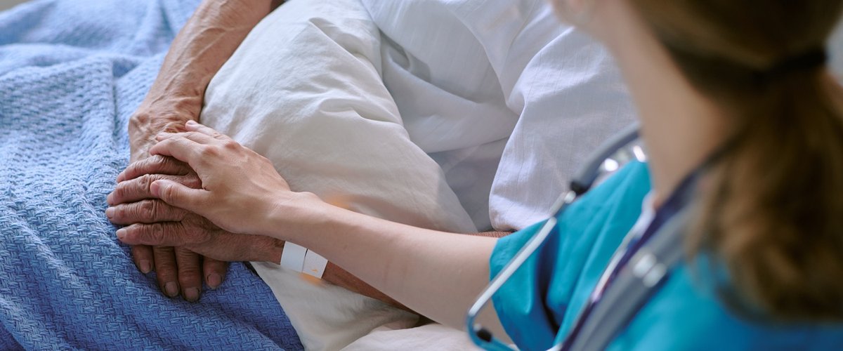 A female healthcare professional with a stethoscope around her neck holds the hand of an older man laying in a hospital bed.