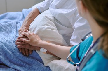 A female healthcare professional with a stethoscope around her neck holds the hand of an older man laying in a hospital bed.