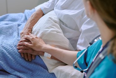 A female healthcare professional with a stethoscope around her neck holds the hand of an older man laying in a hospital bed.