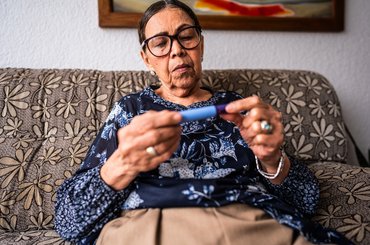 Person sitting on a patterned sofa at home, holding an insulin pen in both hands and checking it closely.