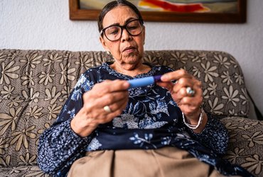 Person sitting on a patterned sofa at home, holding an insulin pen in both hands and checking it closely.