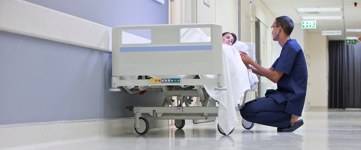 A male nurse kneels next to a female patient laying in a bed on a hospital corridor.