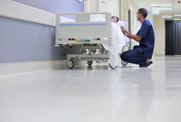 A male nurse kneels next to a female patient laying in a bed on a hospital corridor.