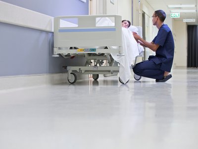A male nurse kneels next to a female patient laying in a bed on a hospital corridor.