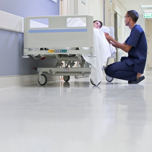 A male nurse kneels next to a female patient laying in a bed on a hospital corridor.
