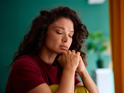 Person sitting indoors with hands clasped under their chin, looking downward in a quiet home setting.