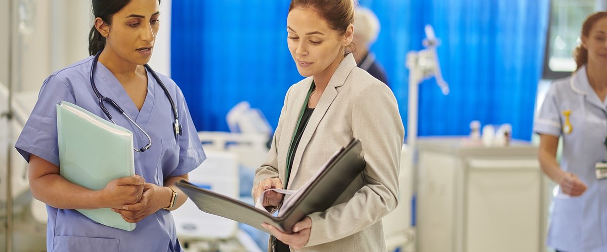A female healthcare professional talks to a female safety investigator on a busy hospital ward.