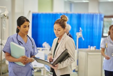 A female healthcare professional talks to a female safety investigator on a busy hospital ward.
