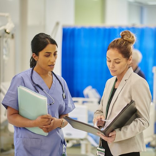 A female healthcare professional talks to a female safety investigator on a busy hospital ward.