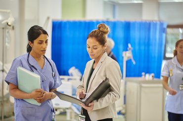 A female healthcare professional talks to a female safety investigator on a busy hospital ward.