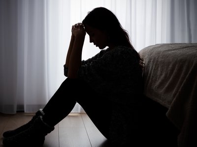 A silhouette of a young woman. She sits on the floor in a dim room with her knees bent, resting her head on her hands.