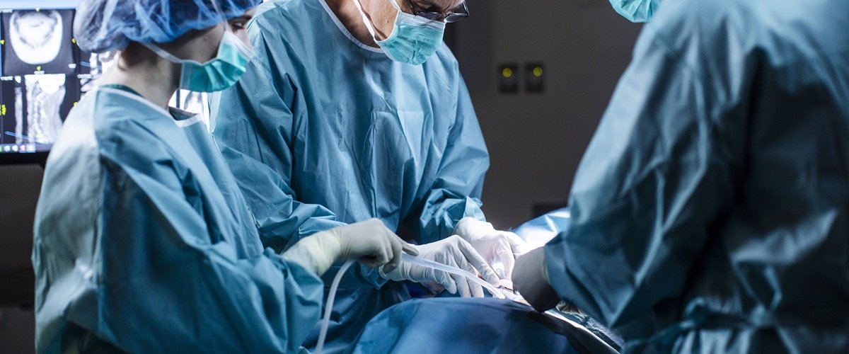 A surgical team wearing protective gowns, masks and caps performs an operation in a hospital operating theatre. Medical staff work around the patient under surgical lighting, with monitoring equipment visible in the background.