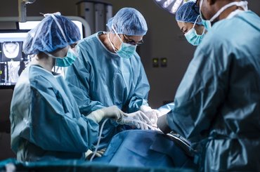 A surgical team wearing protective gowns, masks and caps performs an operation in a hospital operating theatre. Medical staff work around the patient under surgical lighting, with monitoring equipment visible in the background.