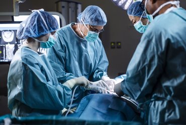 A surgical team wearing protective gowns, masks and caps performs an operation in a hospital operating theatre. Medical staff work around the patient under surgical lighting, with monitoring equipment visible in the background.