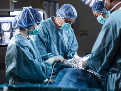 A surgical team wearing protective gowns, masks and caps performs an operation in a hospital operating theatre. Medical staff work around the patient under surgical lighting, with monitoring equipment visible in the background.