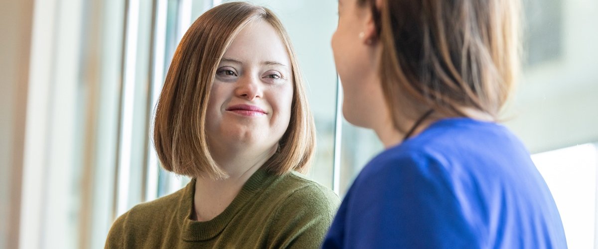 Young person with Down syndrome smiling while talking with a healthcare professional by a window in a hospital setting.