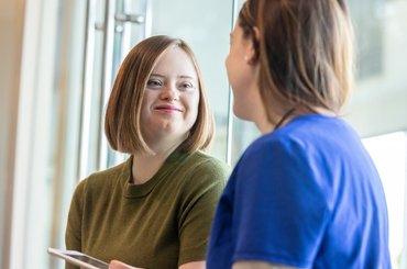 Young person with Down syndrome smiling while talking with a healthcare professional by a window in a hospital setting.