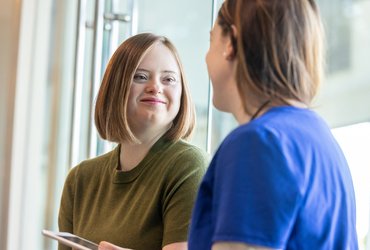 Young person with Down syndrome smiling while talking with a healthcare professional by a window in a hospital setting.