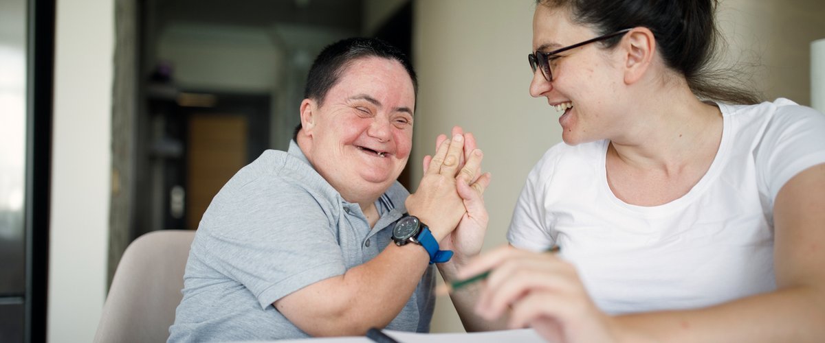 Two people sit together at a table, raising their hands together during a shared activity, with an open notebook in the foreground. One of the people has a learning disability.