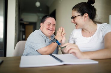 Two people sit together at a table, raising their hands together during a shared activity, with an open notebook in the foreground. One of the people has a learning disability.