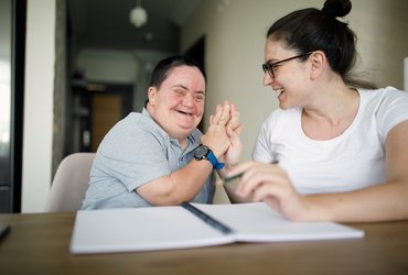 Two people sit together at a table, raising their hands together during a shared activity, with an open notebook in the foreground. One of the people has a learning disability.